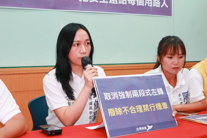 交通安全共同政見記者會，出席者持麥克風發言，前方是「取消強制兩段式左轉 廢除不合理禁行機車」手板